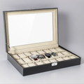 Black 24-slot watch box with glass top open on a grey studio background, cream cushions inside